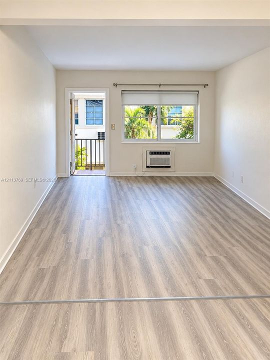 Neutral-toned interior featuring wood-finish flooring, a multi-pane window with roller shade, wall-mounted air conditioning unit, and a private balcony door