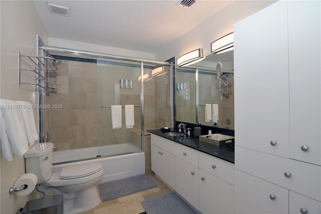 Bathroom featuring a vanity with dark stone countertop, white built-in cabinetry, dual wall-mounted vanity lights, and a tub-shower combination enclosed by a glass door with stone tile surround