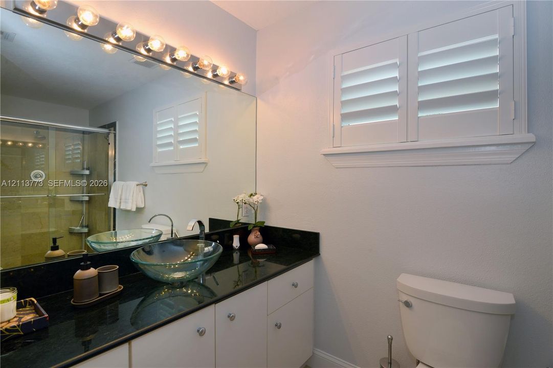 Vanity featuring a dark stone countertop, glass vessel sink, white cabinetry, integrated full-width mirror with multi-bulb light fixture, and plantation shutters