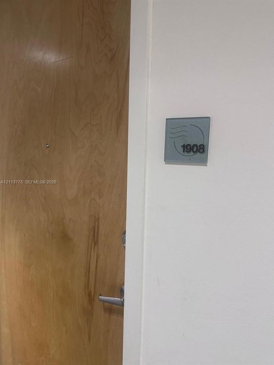 Solid wood-finish door with a metallic peephole and brushed nickel lever handle, accented by a modern wall-mounted unit identifier plaque