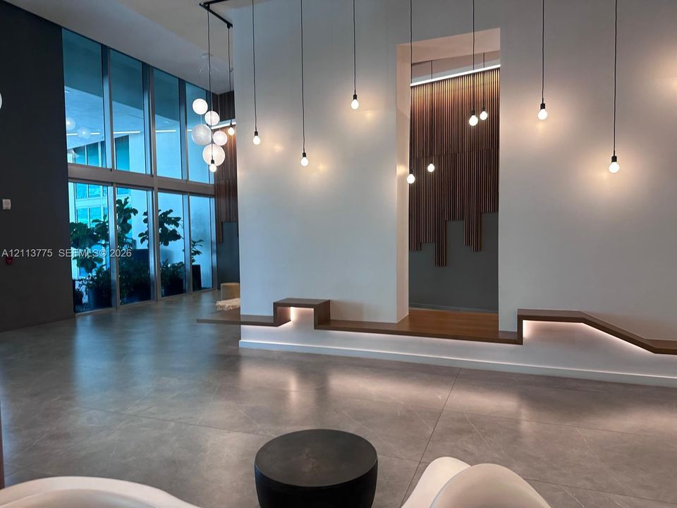 Lobby featuring floor-to-ceiling glass windows, stone-finish flooring, custom wood-slat wall accents, tiered built-in wood benches with integrated LED lighting, and varied pendant lighting fixtures