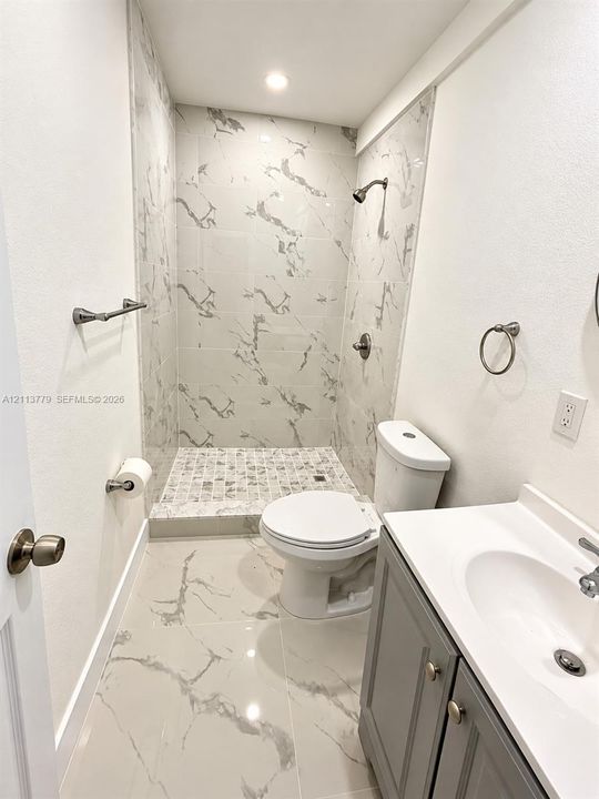 Full bathroom featuring a walk-in shower with stone-look wall tile and pebble-texture floor tile, large-format tile flooring, a gray vanity cabinet with an integrated white sink, and polished nickel hardware