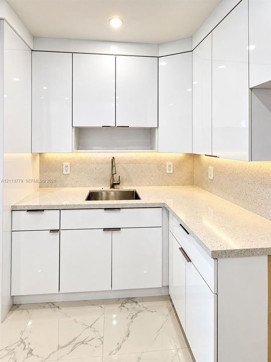 Glossy white cabinetry, stone-composite countertops, stainless steel undermount sink, under-cabinet lighting, and marble-patterned tile flooring