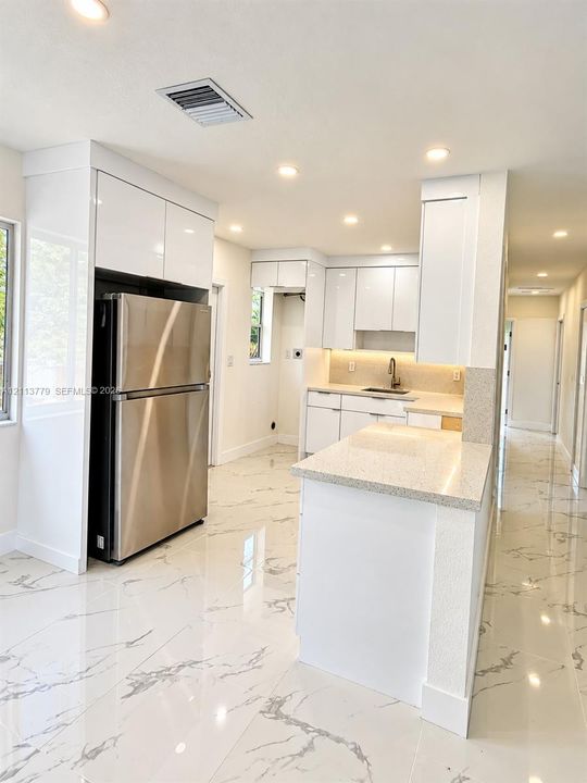 Kitchen featuring white high-gloss cabinetry, stone-finish countertops, stainless steel refrigerator, recessed lighting, and polished stone-finish flooring with veining detail