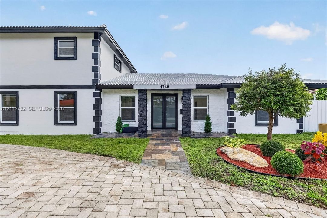 Two-story exterior featuring a white-painted brick facade with dark contrast trim, stone-clad porch columns, a multi-colored stone paver walkway, and a herringbone-patterned concrete paver driveway