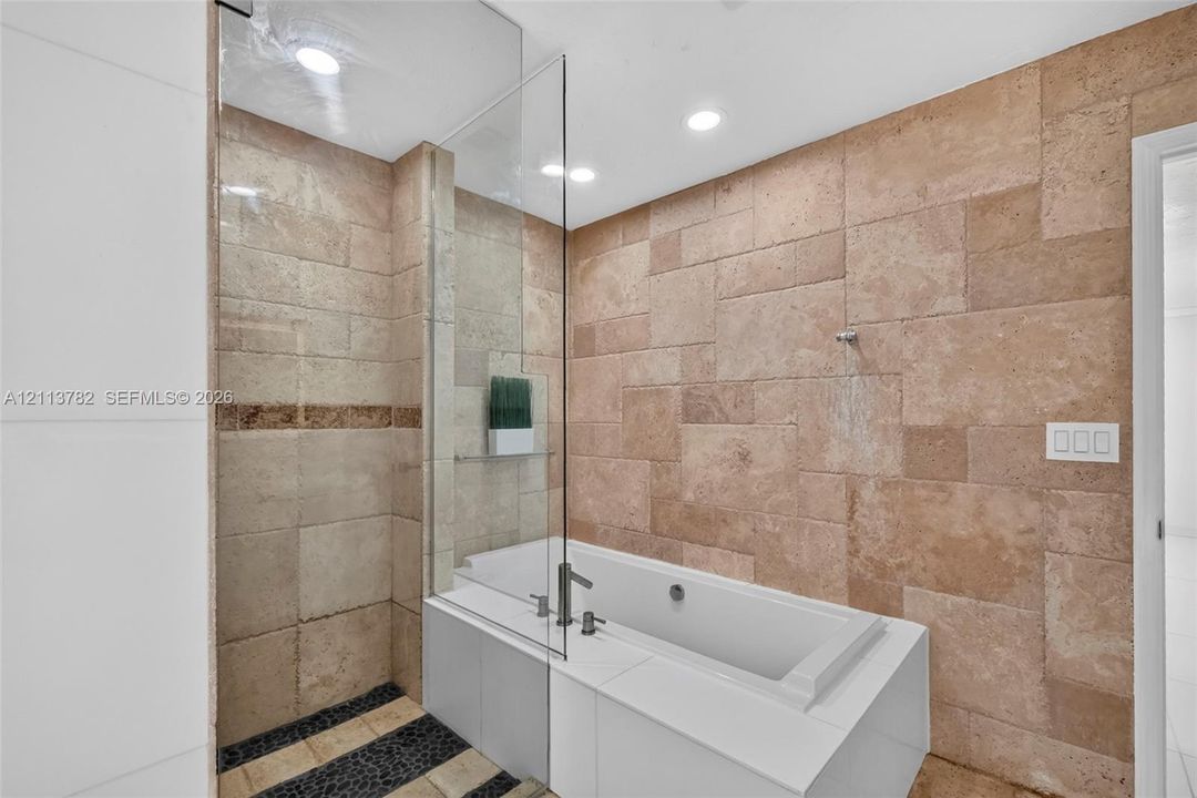 Large-format natural stone wall tiling, integrated soaking tub with a white surround, glass shower partition, recessed ceiling lighting, and pebble-style floor inlay
