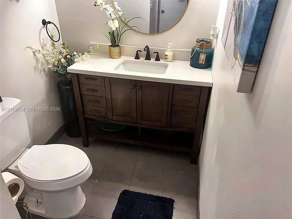 Dark wood vanity with a white composite countertop, rectangular undermount sink, widespread oil-rubbed bronze faucet, circular gold-rimmed mirror, and tiled flooring