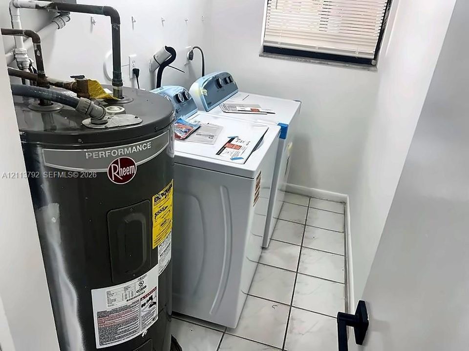 Utility area featuring light-toned tile flooring, a Rheem performance water heater, and a window with horizontal blinds