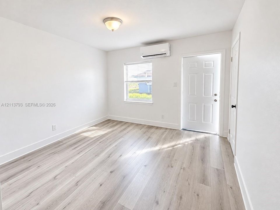 Interior space featuring wood-finish flooring, a wall-mounted ductless mini-split air conditioning unit, a six-panel exterior entry door, and a single-hung window with blinds