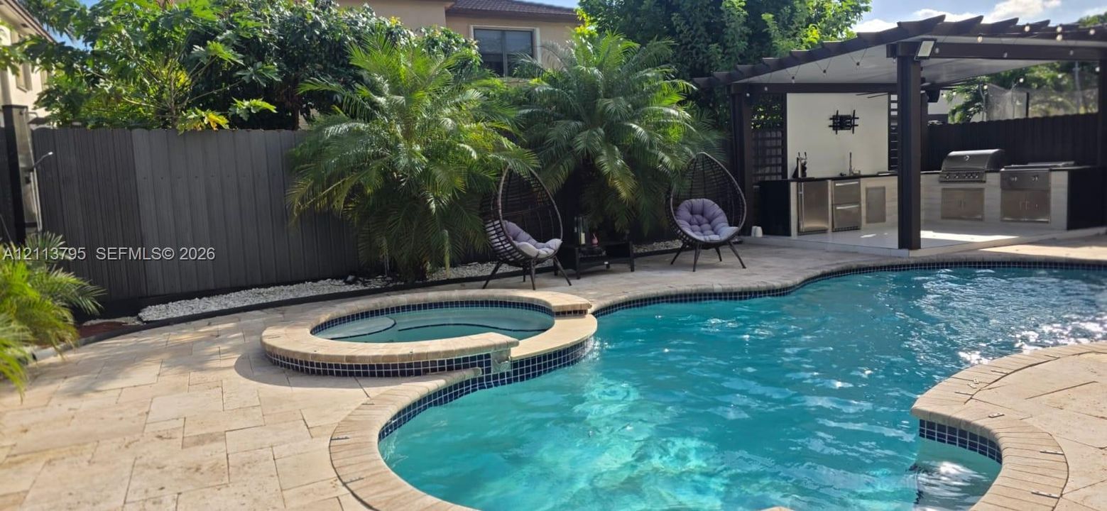 Custom pool with integrated spa, stone-paver decking, covered outdoor kitchen, and privacy fencing