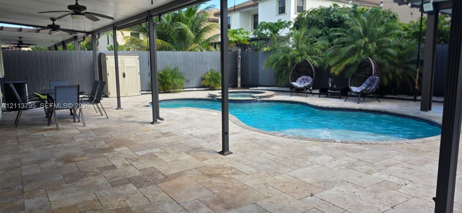 Covered patio featuring stone paver flooring and ceiling fans, leading to an inground swimming pool with an integrated spa and privacy fencing
