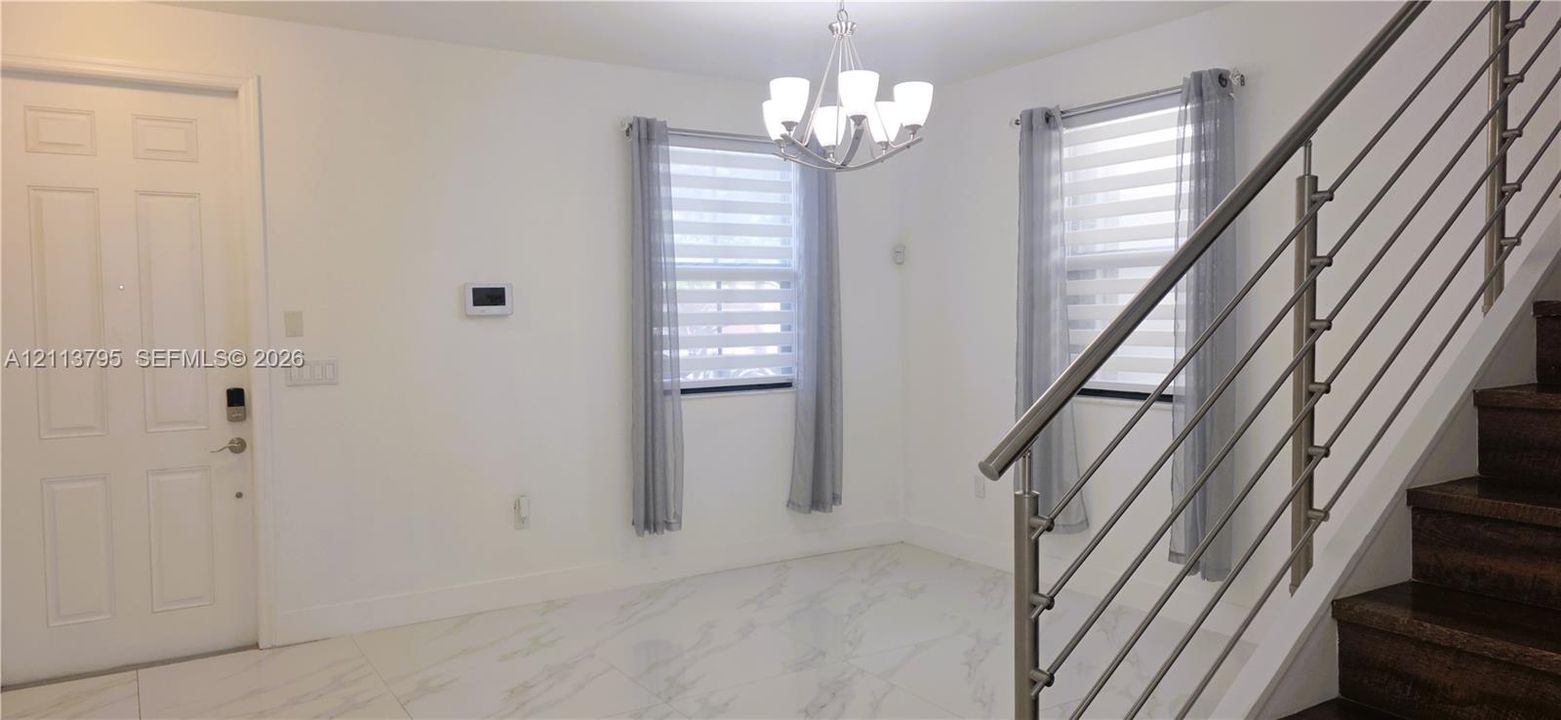 Open-concept interior featuring white tile flooring, a multi-light chandelier, a staircase with metal railings, and dual windows with window treatments