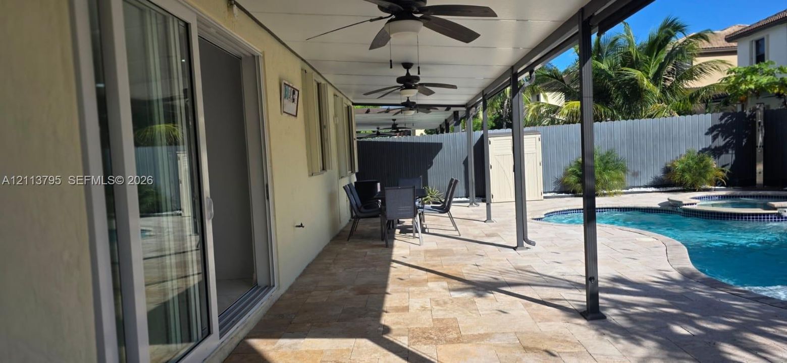 Covered patio featuring stone-tiled flooring, ceiling-mounted fans, sliding glass doors, and a swimming pool with integrated spa