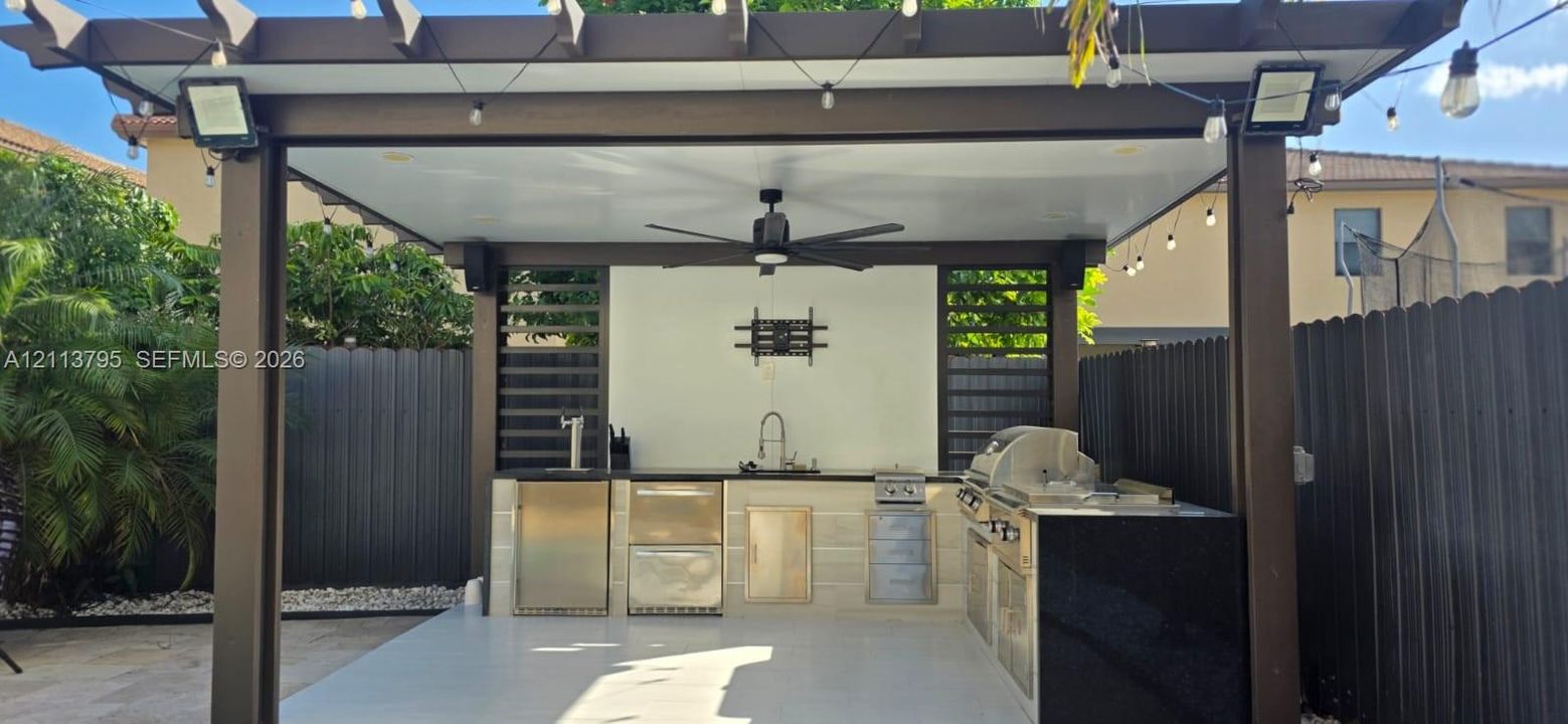 Outdoor kitchen under a covered pergola featuring a stainless steel grill, beverage cooler, sink, solid surface countertops, and a ceiling fan