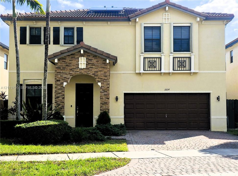 Two-story exterior featuring a stone-veneer entryway, tile roof, decorative wrought-iron balcony railings, and a paved driveway leading to an attached two-car garage