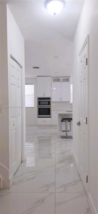 Hallway featuring large-format tile flooring with a marble-finish pattern, white paneled doors, and recessed ceiling lighting, leading toward a kitchen equipped with white cabinetry and built-in stainless steel appliances