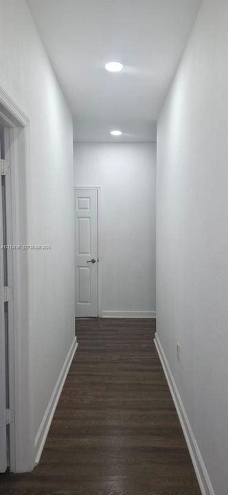 Hallway featuring wood-finish flooring, crisp neutral walls, recessed lighting, and a solid-core panel door with satin nickel hardware