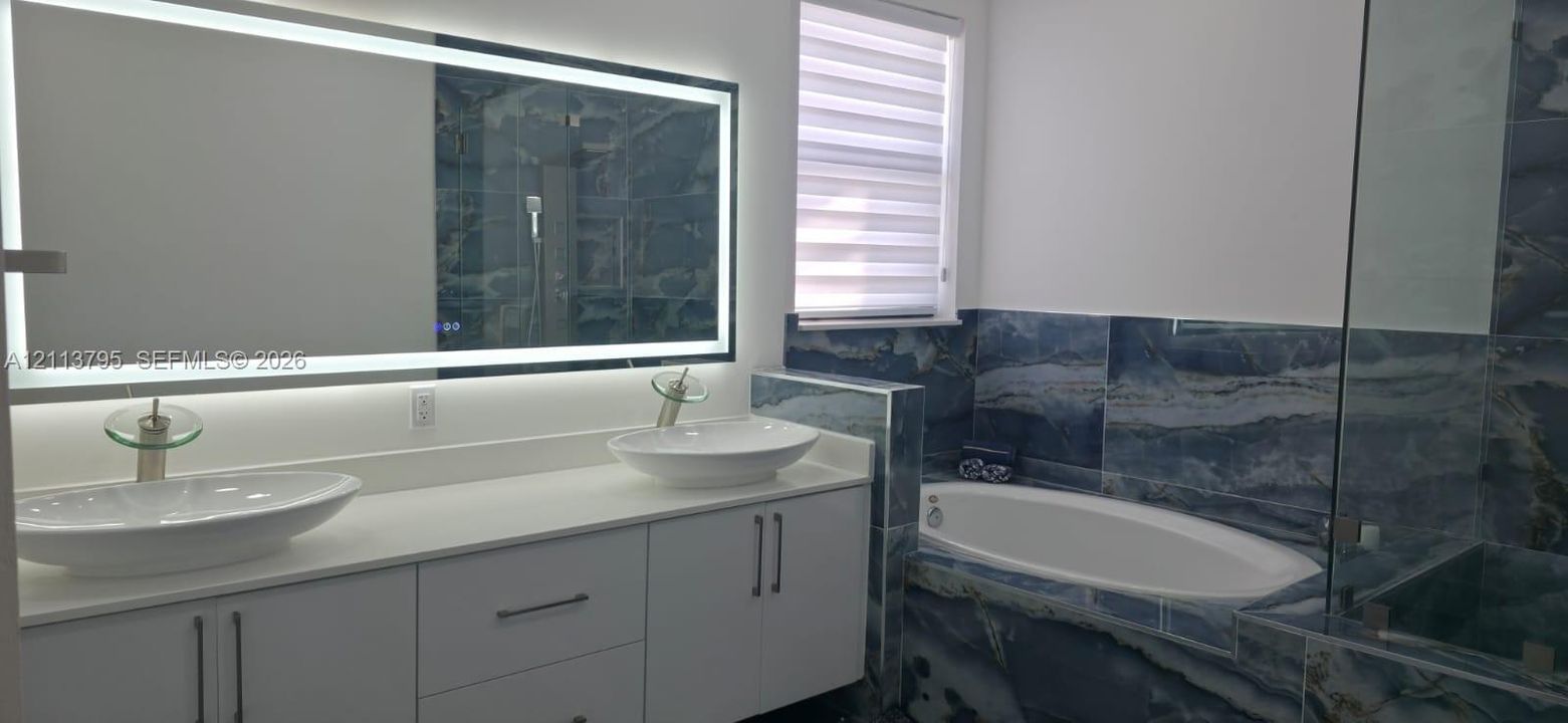 Dual vessel sinks with polished chrome faucets, white cabinetry, solid-surface countertops, expansive LED-backlit mirror, and stone tile surround featuring a deep soaking tub