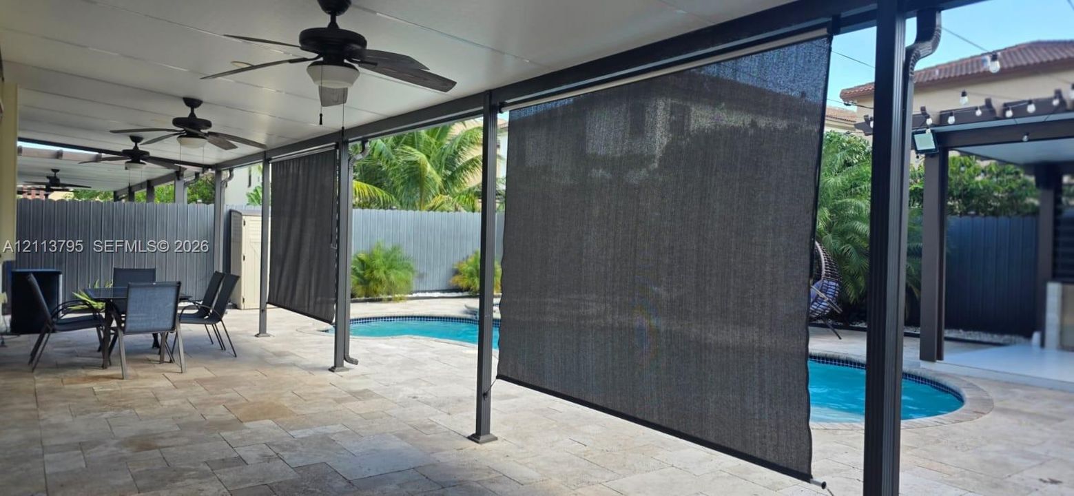 Covered patio featuring stone-tiled flooring, ceiling-mounted fans, retractable solar shades, and an in-ground pool