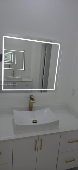 Bathroom vanity featuring a white countertop, rectangular vessel sink, metallic faucet, LED-backlit mirror, and white cabinetry with metallic hardware