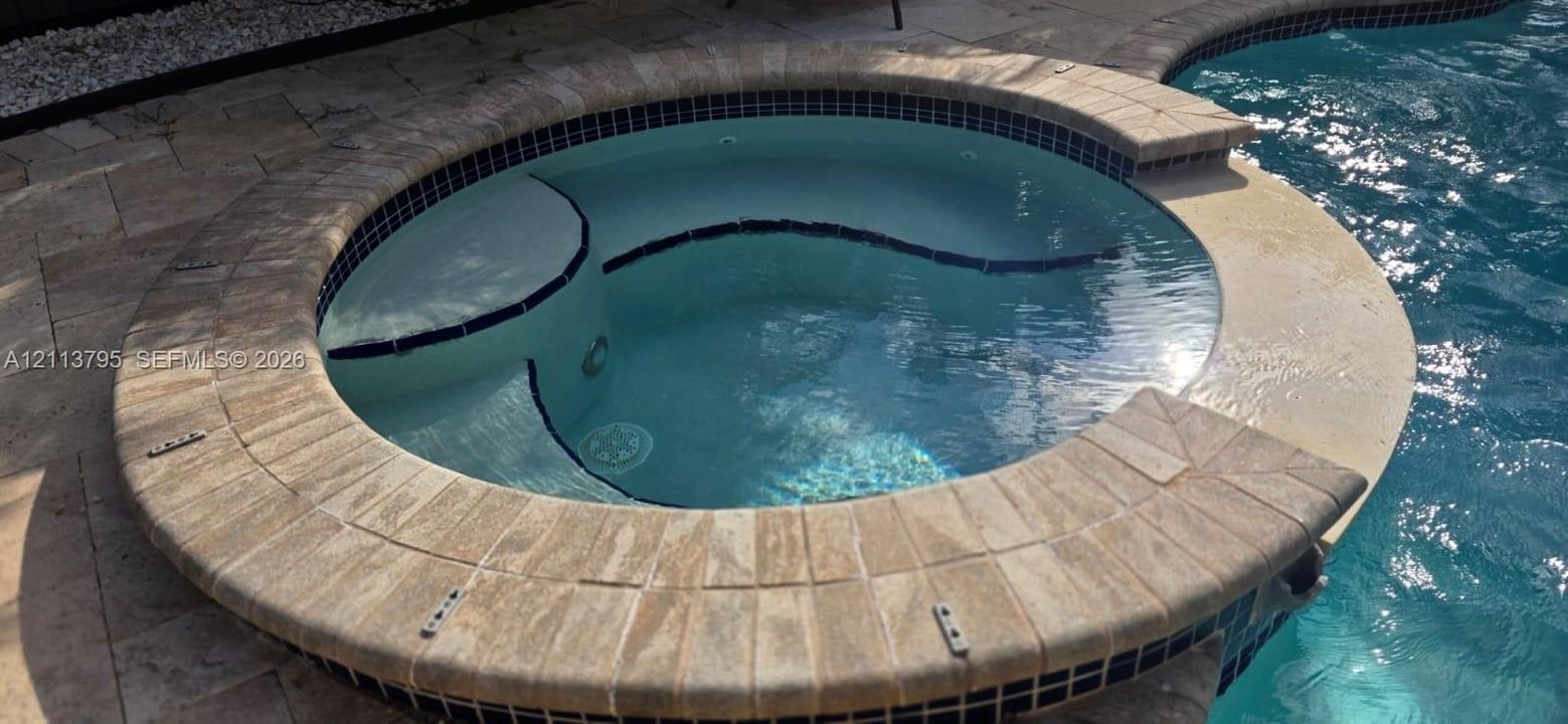 In-ground spa featuring a circular stone-finish coping, decorative tile accents, and integrated steps, set within a stone-paver pool deck
