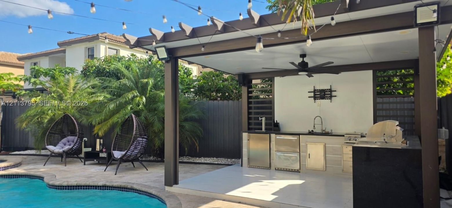 Covered outdoor kitchen featuring stainless steel appliances, a stone-finish facade, a ceiling fan, and privacy screening, situated on a stone-paved patio