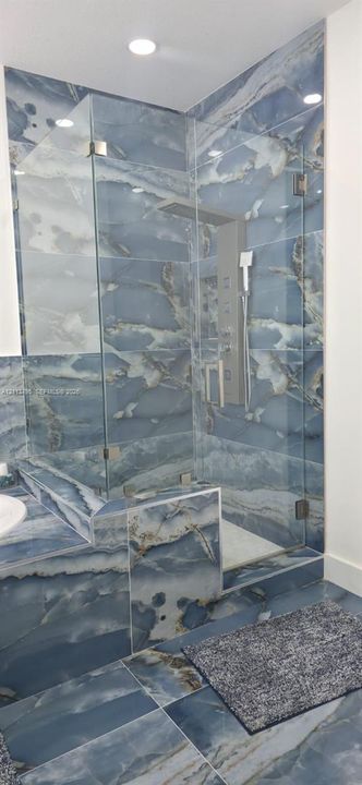 Walk-in shower featuring blue-toned stone-patterned tile, frameless glass enclosure, recessed ceiling lighting, and wall-mounted shower panel system