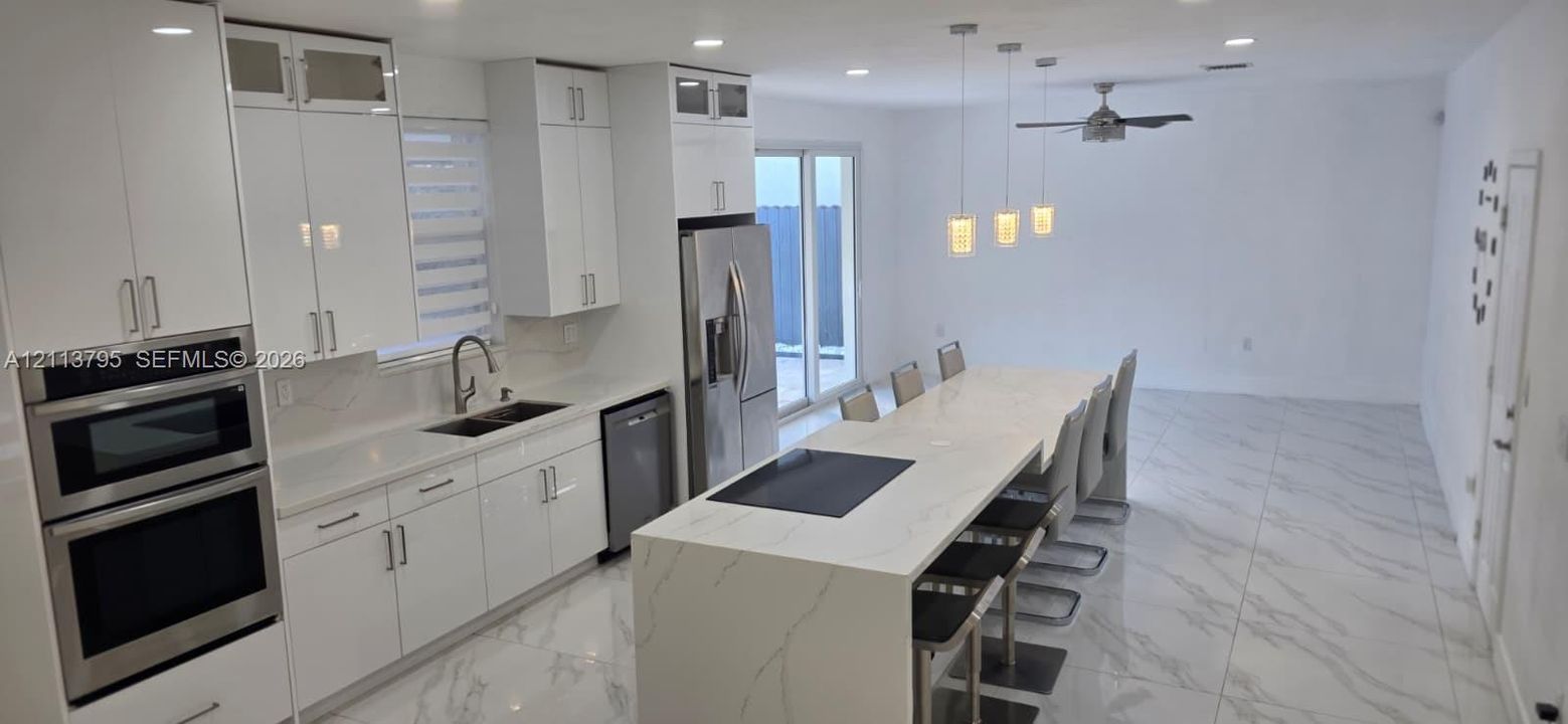 Kitchen featuring high-gloss white cabinetry, expansive stone-finish waterfall island, built-in stainless steel double wall oven, polished stone-finish floor tile, and recessed lighting