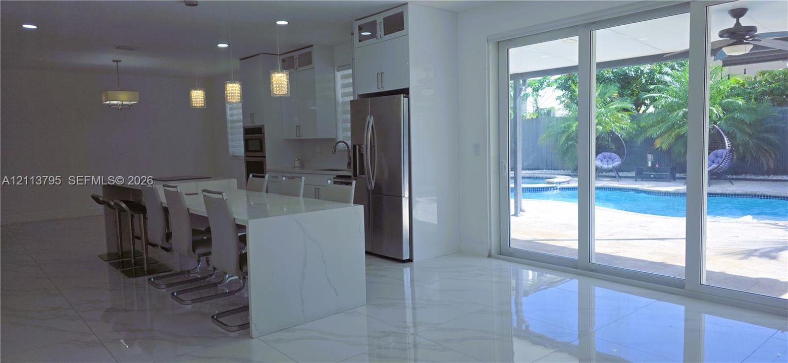 Kitchen featuring high-gloss cabinetry, stone-finish waterfall island, stainless steel appliances, polished tile flooring, and sliding glass doors leading to a pool area