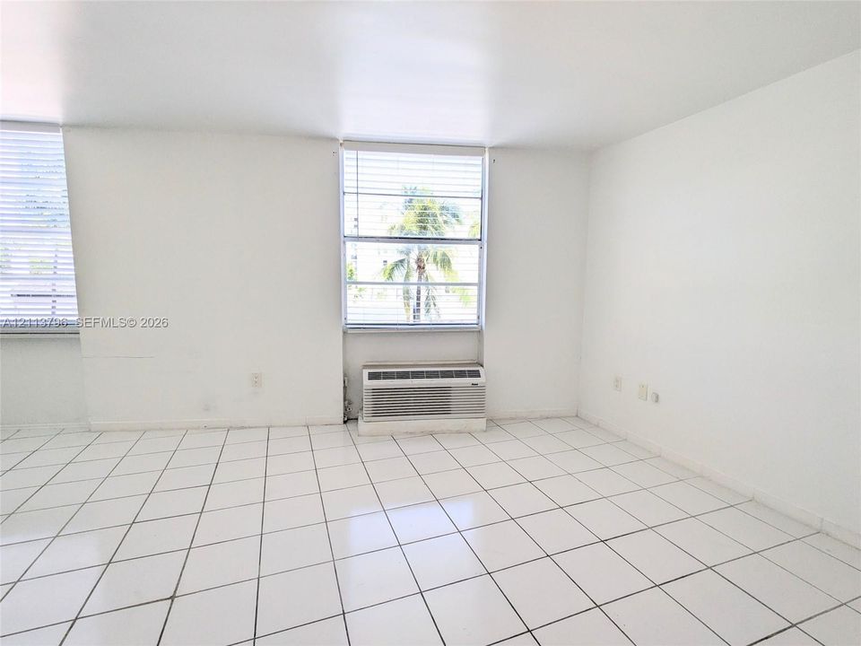 Interior space featuring white tile flooring, neutral walls, dual windows with horizontal blinds, and a built-in wall-mounted climate control unit