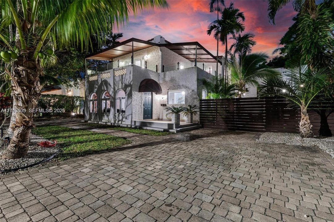 Current photo of house. Two-story exterior featuring textured masonry cladding, arched window detailing, an upper-level covered terrace, and a paved stone driveway
