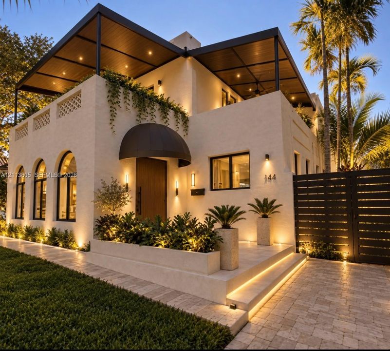 After renovation edited photo of house. Stucco exterior featuring an arched entryway with a metal canopy, upper-level terrace with a covered ceiling, integrated lighting throughout the facade, and stone-paver hardscaping