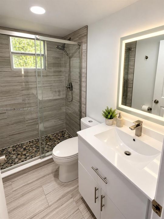 Bathroom featuring a walk-in shower with stone-tile flooring, horizontal wall tile, a glass sliding enclosure, and an integrated LED-lighted mirror over a white vanity