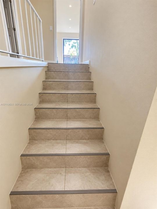 Staircase featuring neutral-tone ceramic tile treads, dark edge nosing, and a white metal handrail