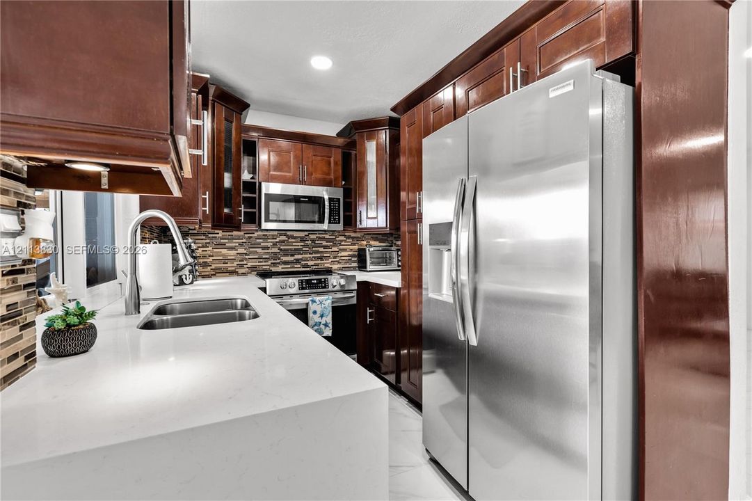 Kitchen featuring stained wood cabinetry, quartz countertops, a multi-tone tile backsplash, stainless steel appliances, and a double-basin undermount sink