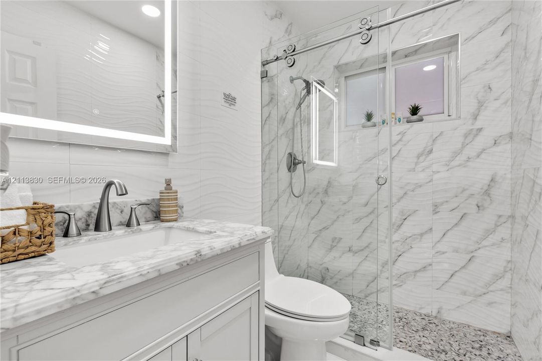 Bathroom featuring a vanity with stone-top basin, integrated LED-backlit mirror, wall-mounted faucet, and glass-enclosed shower with tiled surround and textured flooring