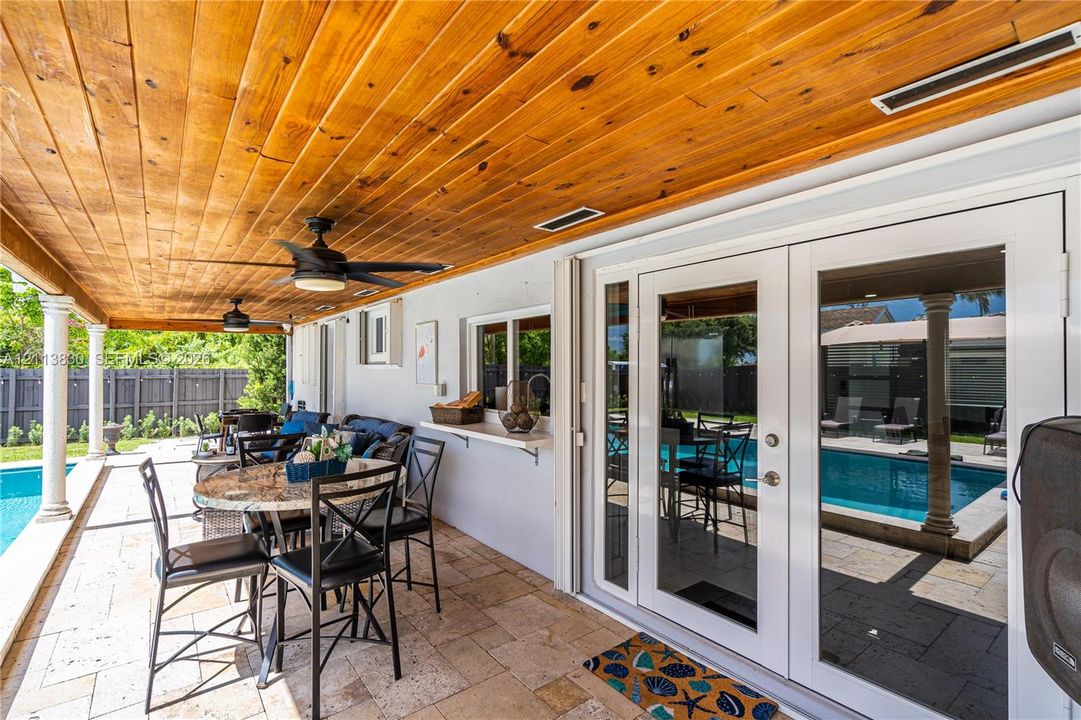 Covered patio featuring a wood-plank ceiling, natural stone tile flooring, ceiling fans, built-in pass-through service shelf, and French doors