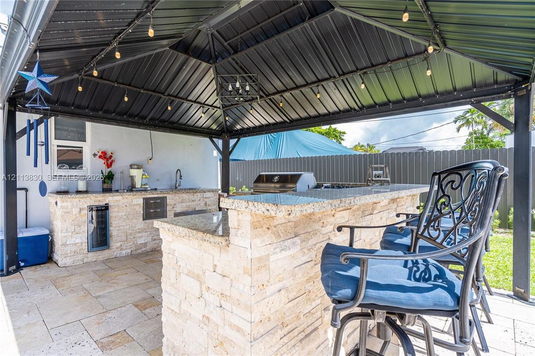 Covered outdoor kitchen featuring a stone-clad bar, natural stone countertop, built-in beverage refrigerator, and metal gazebo structure
