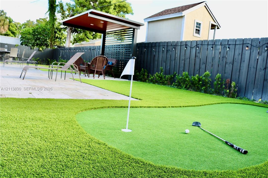 Outdoor putting green featuring synthetic turf, paver patio, covered pavilion with slatted privacy screen, and fenced perimeter