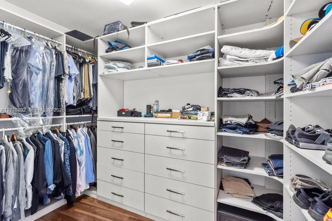 Custom walk-in closet featuring white built-in shelving, floor-to-ceiling storage, a ten-drawer dresser unit with metallic pulls, and wood-finish flooring