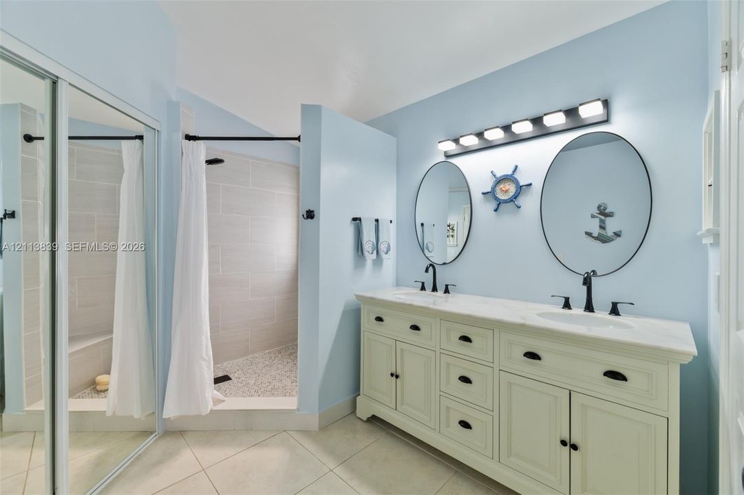Double vanity featuring a stone-top counter, dual oval mirrors, matte black hardware, and a walk-in shower with tile surround