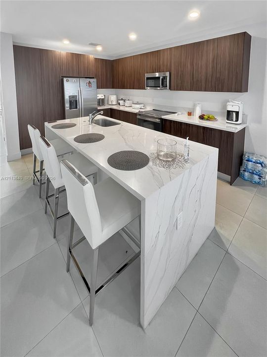 Modern kitchen featuring a waterfall-edge island with a stone-finish countertop, flat-panel wood-finish cabinetry, stainless steel appliances, and large-format tile flooring