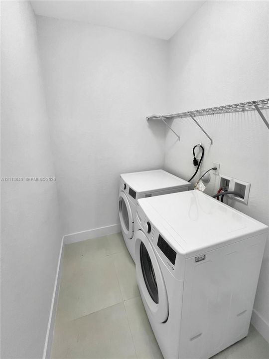 Laundry space featuring large-format tile flooring, white walls, and a wall-mounted wire storage shelf