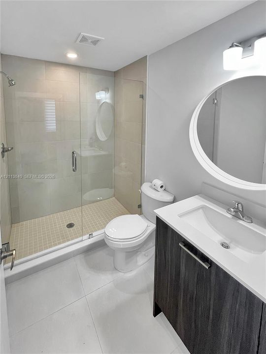 Bathroom featuring a glass-enclosed walk-in shower with tile surround, vanity with wood-finish cabinetry, white countertop with integrated sink, and an oval wall mirror