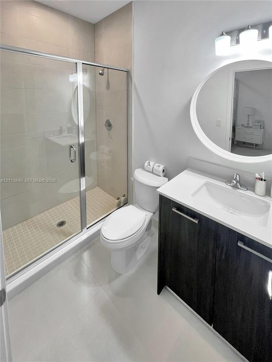 Bathroom featuring a vanity with dark-finish cabinetry, a white countertop with an integrated sink, a walk-in shower with a glass enclosure, and a circular wall-mounted mirror