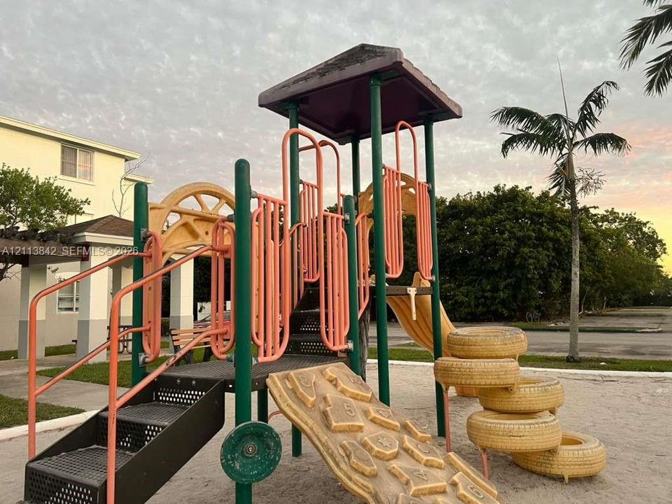 Outdoor recreational structure featuring a multi-level platform, metal stair access, two slides, and a stacked tire climbing component situated on a sand-surfaced area