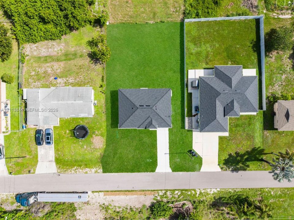 Aerial perspective of a property featuring a multi-pitched shingled roof, concrete driveway, and expansive grass lot enclosed by perimeter fencing