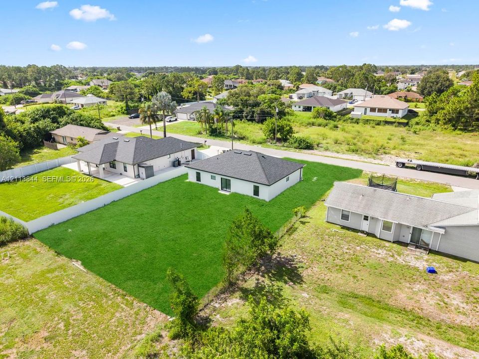 Aerial perspective featuring a single-story residence with a dark shingled roof, light-toned exterior finish, and a grass-covered backyard enclosed by a white privacy fence