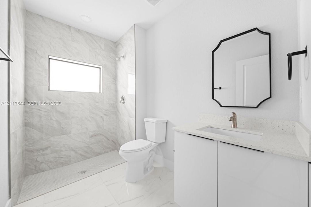 Full bathroom featuring a walk-in shower with stone-finish tile surround, white vanity cabinet with quartz countertop, undermount sink, and geometric wall-mounted mirror