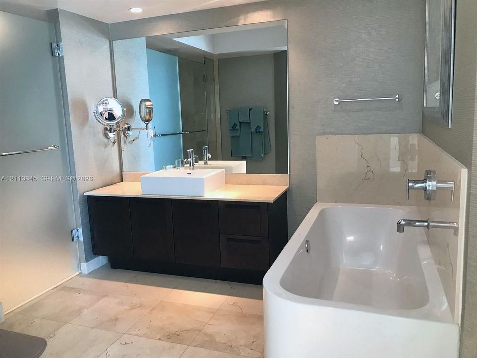 Dark wood-finish vanity with a rectangular vessel sink and countertop, large wall-mounted mirror, soaking tub with polished chrome fixtures, and large-format stone-finish flooring